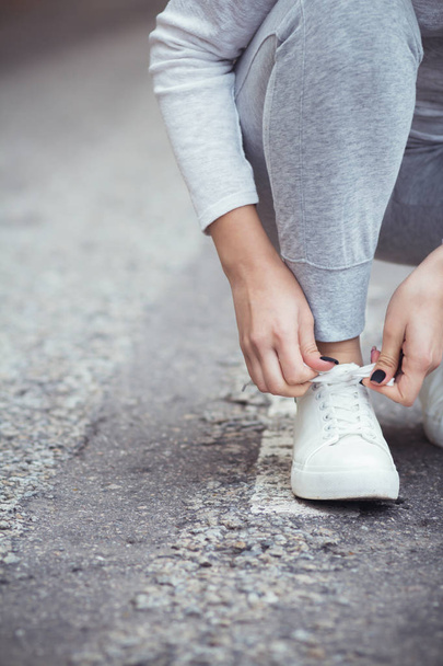 girl squatted down to tie shoelaces on white sneakers on asphalt road, autumn sport concept outdoors - Photo, Image