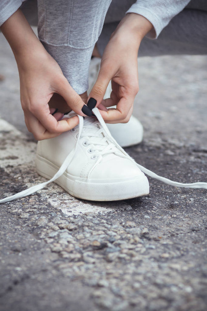 girl squatted down to tie shoelaces on white sneakers on asphalt road, autumn sport concept outdoors - Фото, изображение