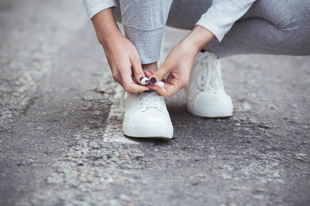 girl squatted down to tie shoelaces on white sneakers on asphalt road, autumn sport concept outdoors - Foto, afbeelding