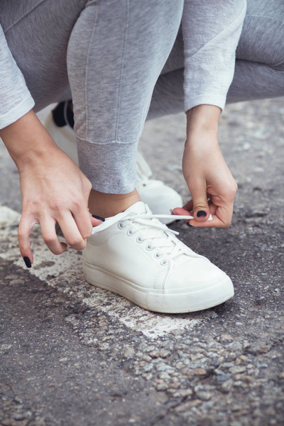 girl squatted down to tie shoelaces on white sneakers on asphalt road, autumn sport concept outdoors - Photo, image