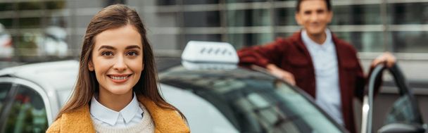Enfoque selectivo de la mujer sonriendo a la cámara con el taxista en el fondo, plano panorámico
  - Foto, Imagen