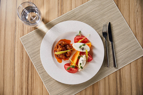 top view of delicious restaurant dish with eggplant caviar and tomatoes served on wooden table with water and cutlery - Fotografie, Obrázek