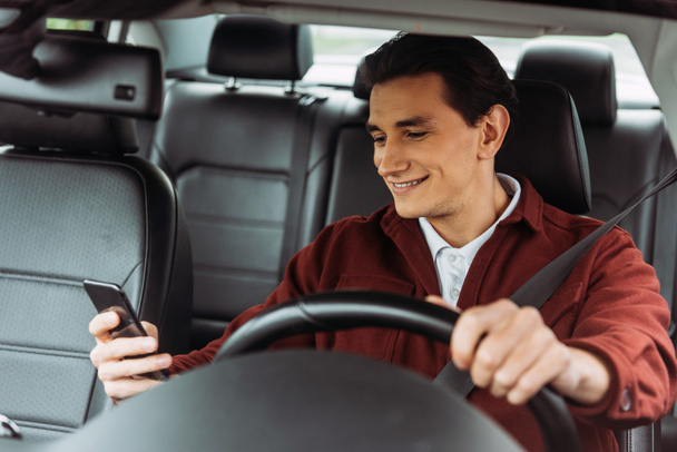 Smiling man looking at smartphone while driving car - Фото, изображение