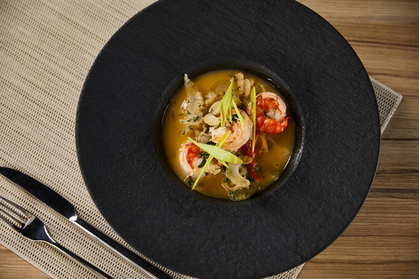 top view of delicious restaurant soup with shrimps in black plate served on napkin with cutlery - Fotografie, Obrázek