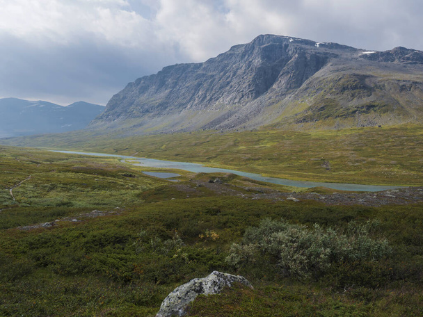 Beautiful wild Lapland nature landscape with blue glacial river, birch tree bushes, snow capped mountains and dramatic clouds. Northern Sweden summer at Kungsleden hiking trail. - Φωτογραφία, εικόνα
