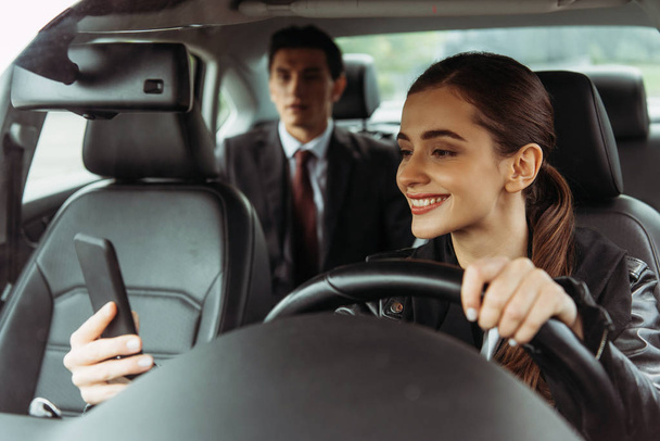 Uśmiechnięty taksówkarz korzystający ze smartfona z biznesmenem w tle - Zdjęcie, obraz