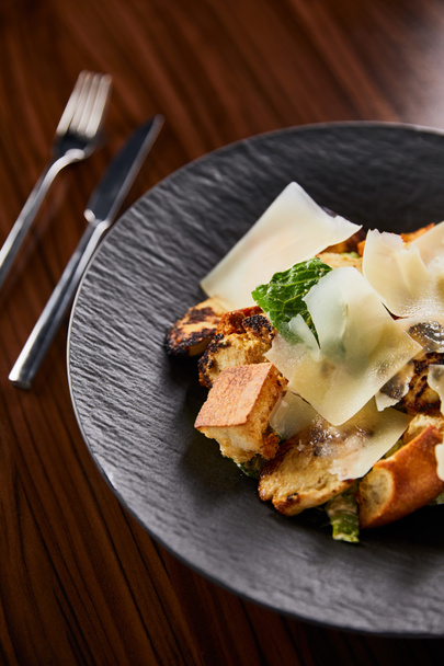 selective focus of tasty Caesar salad with Parmesan slices served on plate with cutlery on wooden table - Foto, imagen