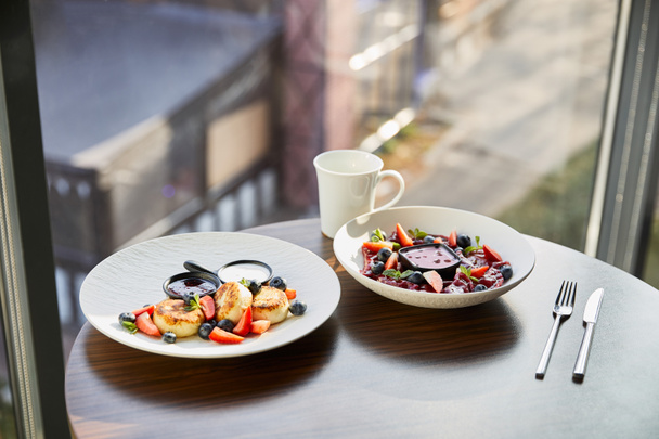 традиционные украинские вареники и сирники с ягодами подаются в белой тарелке с соусом возле столовых приборов и кубком на деревянном столе в ресторане
 - Фото, изображение