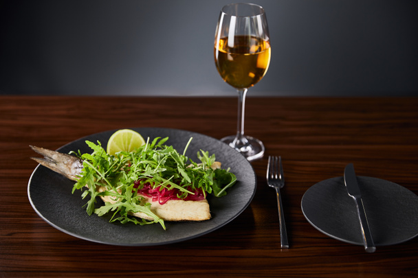 tasty restaurant fish steak with lime and arugula on wooden table near cutlery and white wine on black background - Fotó, kép