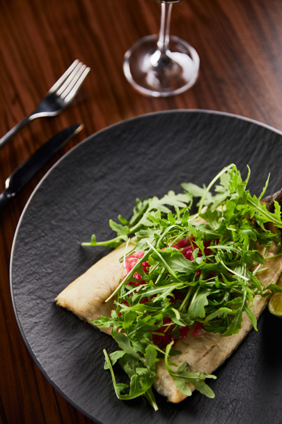 selective focus of tasty restaurant fish steak with lime and arugula on wooden table near cutlery - Foto, imagen