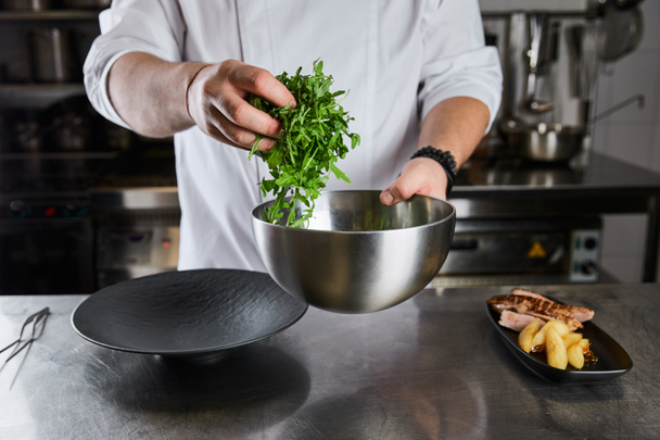 cropped view of chef cooking dish with arugula, meat and potato at kitchen in restaurant - Photo, Image
