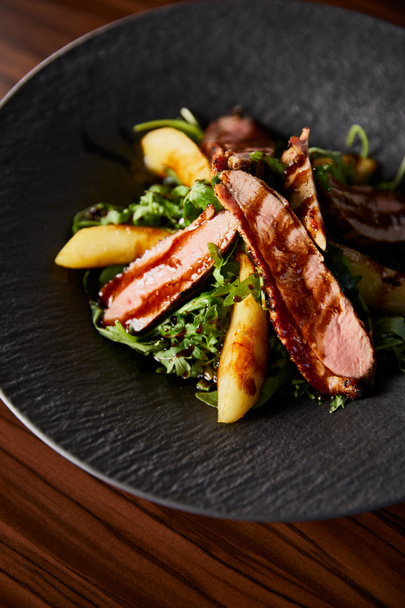 delicious warm salad with arugula, potato and meat in black plate on wooden table - Photo, Image