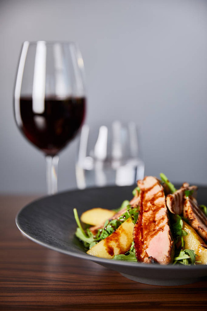 foyer sélectif de délicieuse salade chaude avec roquette, pomme de terre et viande en assiette noire sur table en bois près du vin rouge, eau sur fond gris
 - Photo, image