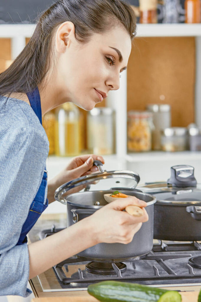 Beautiful girl is tasting food and smiling while cooking in kitchen at home - 写真・画像