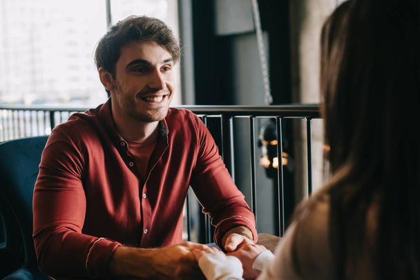 χαμογελαστός νεαρός άνδρας κοιτάζοντας φίλη και κρατώντας τα χέρια της στο μπαλκόνι στην καφετέρια - Φωτογραφία, εικόνα