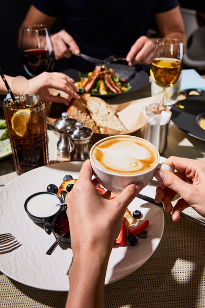 abgeschnittene Ansicht einer Frau, die köstliche Syrniki isst und beim Abendessen mit Freunden im Restaurant Cappuccino trinkt  - Foto, Bild