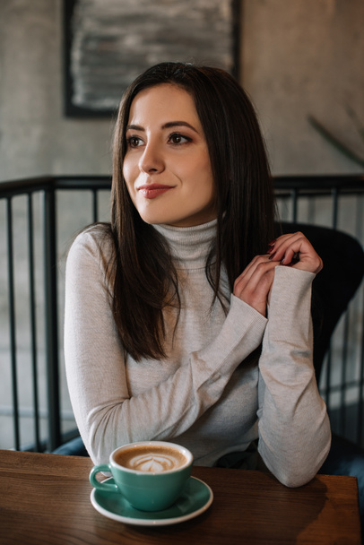young woman sitting at wooden table with cappuccino on balcony in coffee shop - Photo, Image
