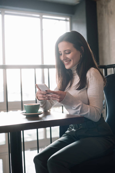 smiling young woman sitting at wooden table with cappuccino and using smartphone on balcony in coffee shop - Foto, Imagem