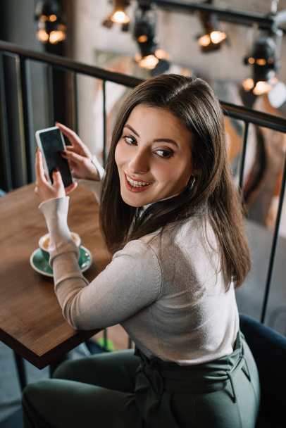 smiling young woman sitting at wooden table with cappuccino and using smartphone on balcony in coffee shop - Foto, Imagen
