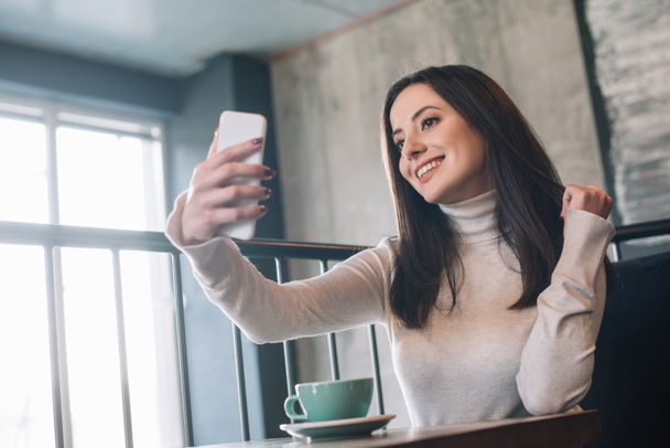 笑顔の若い女性がカプチーノと木製のテーブルに座ってコーヒーショップのバルコニーでスマートフォンで自撮り - 写真・画像