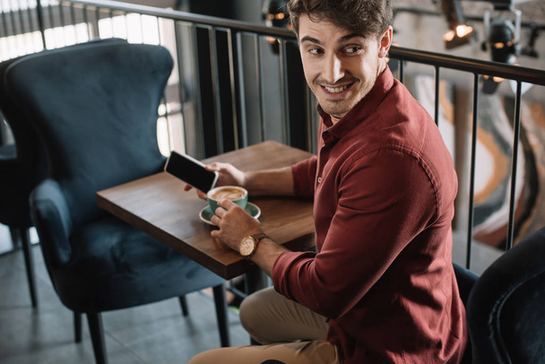 uśmiechnięty młody człowiek siedzi przy drewnianym stole z cappuccino i trzyma smartfona na balkonie w kawiarni - Zdjęcie, obraz