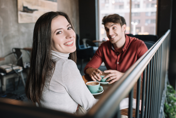 selective focus of smiling girl looking at camera while drinking coffee with man on balcony in coffee shop - Photo, Image