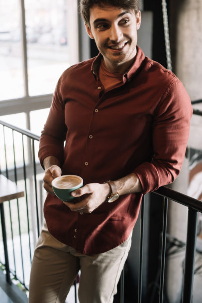 smiling man standing on balcony with cappuccino in coffee shop - Фото, изображение