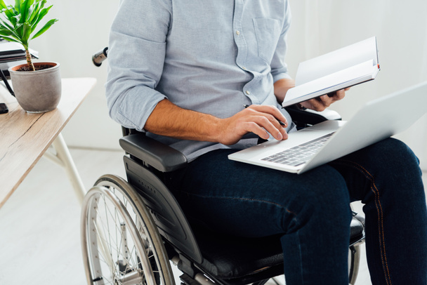 Cropped view of man in wheelchair holding notebook and laptop - Foto, afbeelding