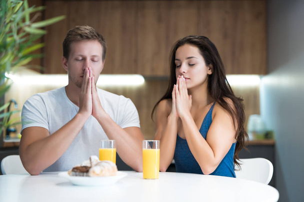 young couple saying prayer before start eating their breakfast in the kitchen family traditions - Φωτογραφία, εικόνα