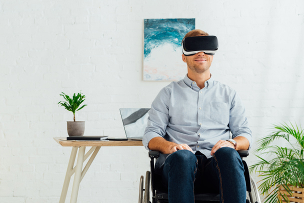 Uśmiechnięty niepełnosprawny mężczyzna na wózku inwalidzkim korzystający z zestawu słuchawkowego wirtualnej rzeczywistości w salonie - Zdjęcie, obraz