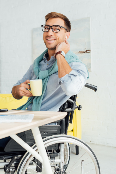 Tekerlekli sandalyedeki gülen adam kablosuz kulaklık kullanıyor ve elinde çay fincanı tutuyor. - Fotoğraf, Görsel