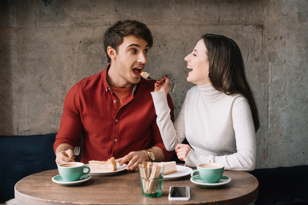 happy girlfriend feeding boyfriend with cheesecake in coffee shop - Photo, Image