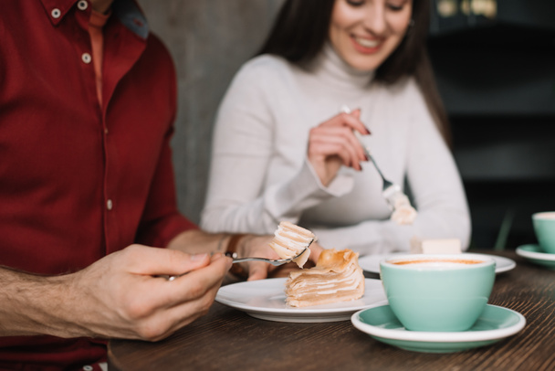 przycięty widok szczęśliwej pary jedzącej sernik i pijącej cappuccino w kawiarni - Zdjęcie, obraz