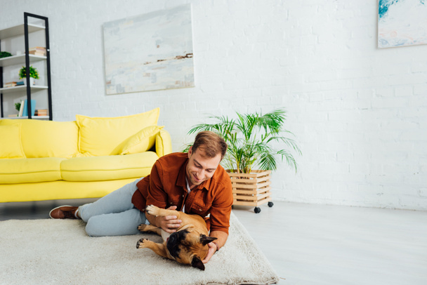 Smiling man playing with french bulldog on floor in living room - Foto, Bild