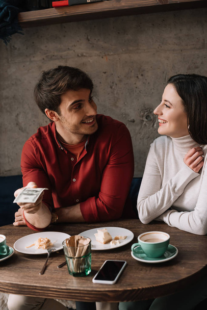happy couple looking at each other while man holding money in coffee shop - Photo, Image