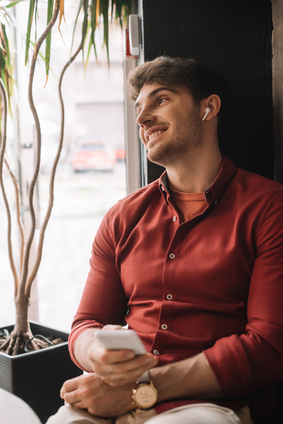 smiling man listening music in wireless earphones and holding smartphone smartphone near window - Zdjęcie, obraz