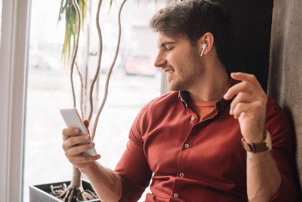 smiling man listening music in wireless earphones and using smartphone smartphone near window - Photo, image