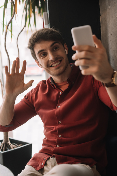 Lächelnder Mann mit drahtlosen Kopfhörern, der einen Videochat auf dem Smartphone nutzt und vor dem Fenster mit der Hand winkt - Foto, Bild