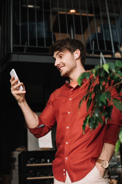 uśmiechnięty mężczyzna w słuchawkach bezprzewodowych za pomocą smartfona w pobliżu zielonej rośliny - Zdjęcie, obraz