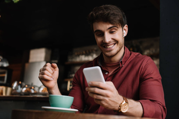 χαρούμενος άνθρωπος σε ασύρματα ακουστικά χρησιμοποιώντας smartphone στο τραπέζι με φλιτζάνι καφέ - Φωτογραφία, εικόνα