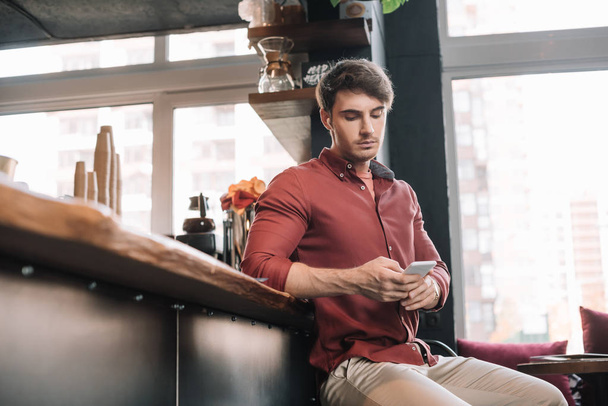 handsome man sitting near bar counter in wireless earphones using smartphone - Foto, Imagem