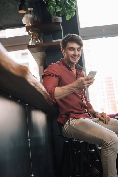 sourire bel homme assis près du comptoir de bar dans les écouteurs sans fil à l'aide d'un smartphone
 - Photo, image