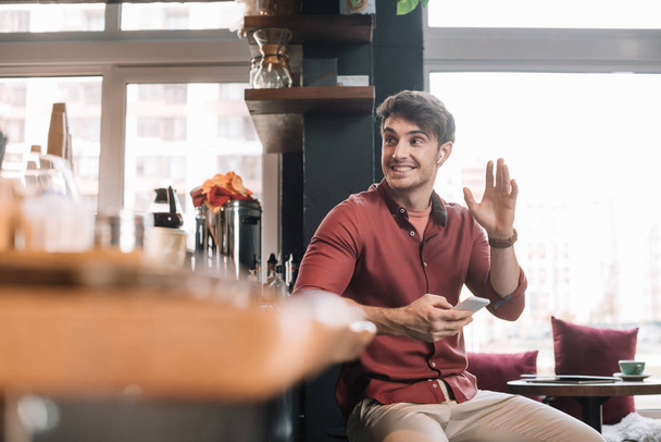 smiling handsome man sitting near bar counter in wireless earphones with smartphone and waving hand - Foto, Imagen