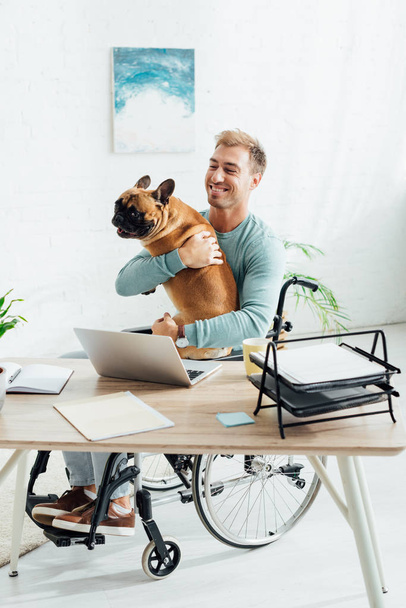 Smiling man in wheelchair holding french bulldog at home - Foto, imagen