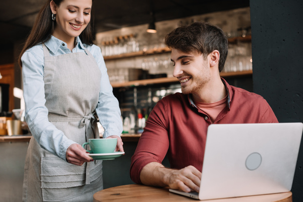 smiling waitress serving coffee to freelancer with laptop - Photo, Image