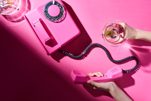 Cropped view of woman holding cocktail and telephone handset beside astray with cigarette butts on pink surface - Fotografie, Obrázek