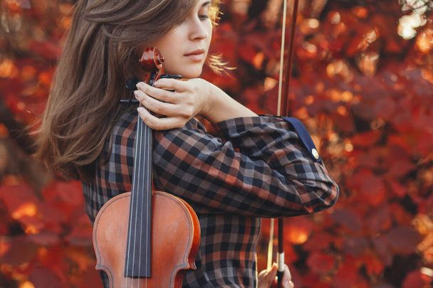 young woman with red violin and and bow on a background of autumn nature, romantic girl engaged in musical art, a concept of hobby, concert outdoors - 写真・画像