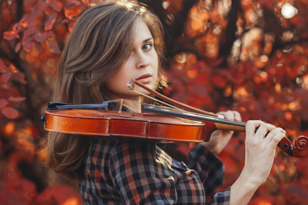 young woman with red violin and and bow on a background of autumn nature, romantic girl engaged in musical art, a concept of hobby, concert outdoors - Foto, Bild