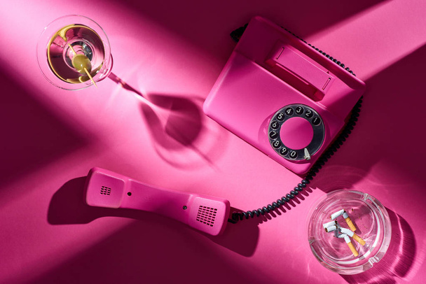 Top view of pink telephone, cocktail and astray with cigarette butts on pink surface - Фото, зображення