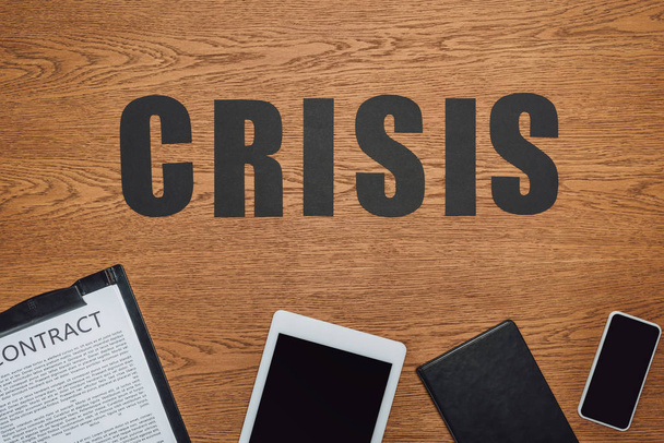top view of word crisis near smartphone, digital tablet, notebook and clipboard with contract on wooden desk - Foto, Imagem